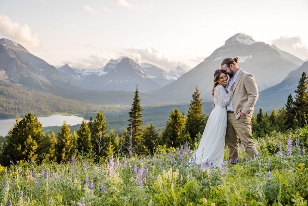 Bride and Groom hugging in a field of wildflowers, including lupine, during an elopement in Glacier National Park with mountains behind them.