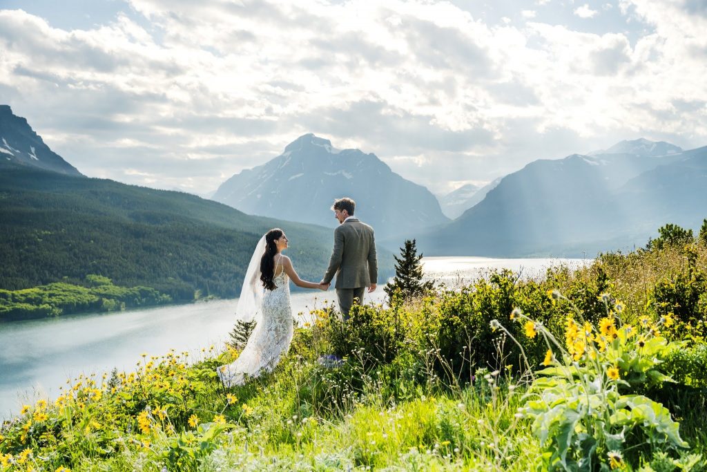 Bride and Groom holding hands in a field of Balsamroot wildflowers with mountains and water in front of them.