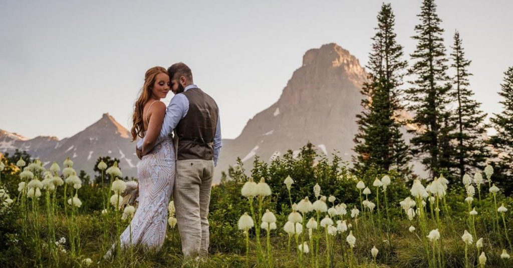 Bride and Groom hugging in a field of Beargrass wildflowers, during an elopement in Glacier National Park with mountains behind them.