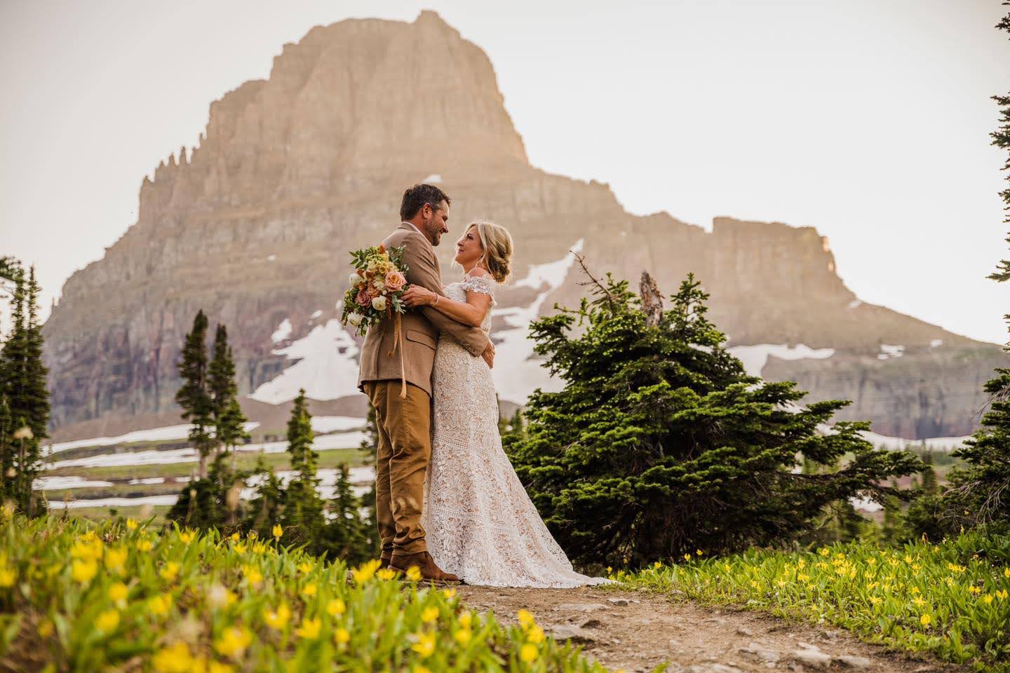 Bride and Groom hugging during a Glacier National Park elopement, surrounded by Glacier Lily, with mountains behind them.