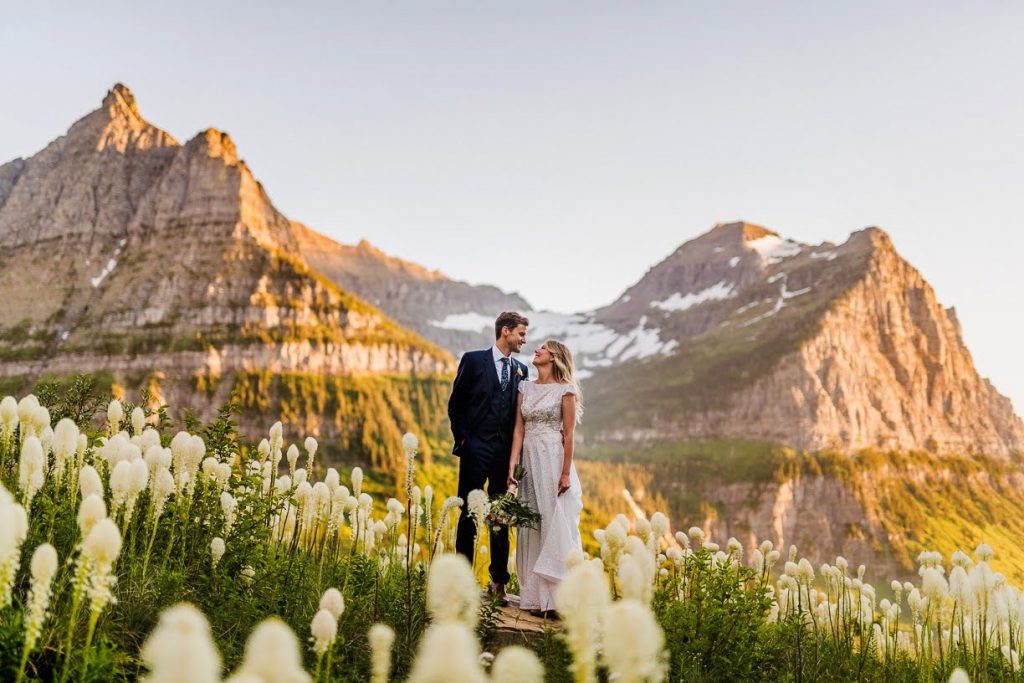 Bride and Groom hugging in a field of Beargrass wildflowers, during an elopement in Glacier National Park with mountains behind them.