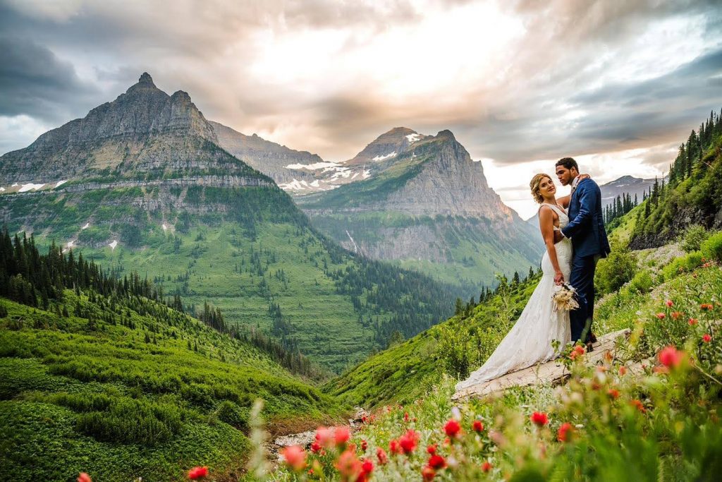 Bride and Groom hugging in a field of wildflowers, including Indian Paintbrush, during an elopement in Glacier National Park with mountains behind them.