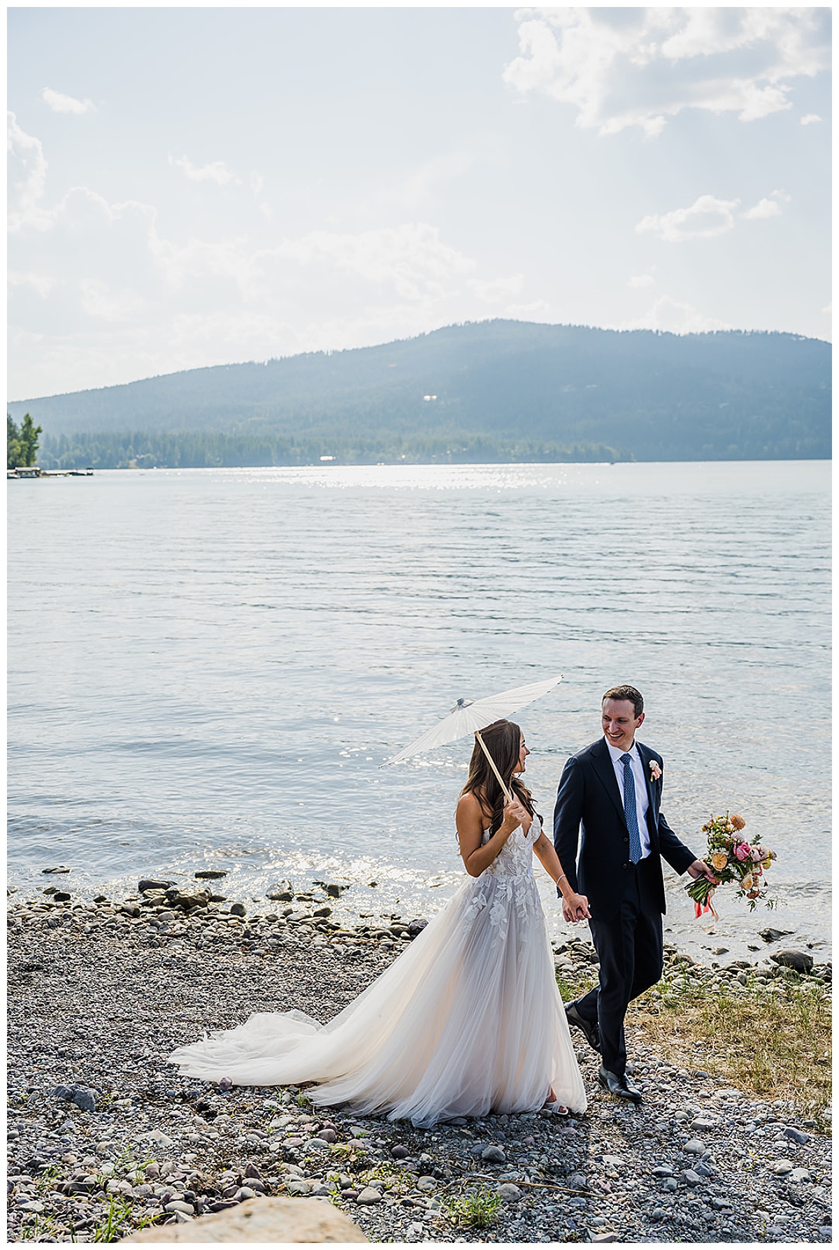 Bride and Groom walking on the rocky shoreline of Whitefish Lake after their wedding ceremony. Bride is holding a parasol and Groom is holding her bouquet of flowers.